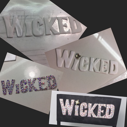 I started the sign by tracing out the letters onto card and then inserted that into a poly pocket.\nUsing white acrylic, I made the letters and then refined them with a hand file before coating them in a matte top coat.\nTaking AB Crystals in SS5, SS7 and SS9, I secured them with hard gel. This was to signify the bright lit up sign outside of the theatre.\nThe Luminous Flame crystals were a perfect finishing touch above the “i”.\nI mounted the letters onto a black ground which I made from the same black acrylic powder as Elphaba.\nTo make my sign stand up, I used thin wooden sticks which I painted black.\nNOT PICTURED: I made a monomer mix of green liquid and a green iridescent pigment and created clusters of “Emeralds” at the base of each post to give some added dimension.